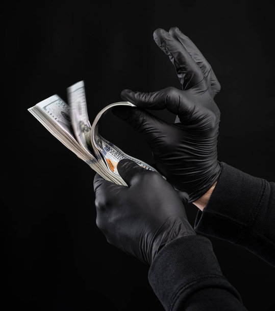Finances concept. Man in black gloves holding pack of dollars over black background - Photo, Image