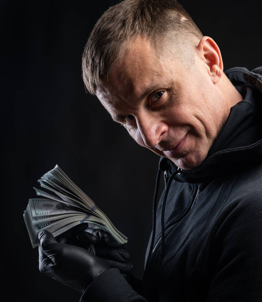 Business and finances concept. Man in black hoody holding pack of american dollars over black background - Photo, Image