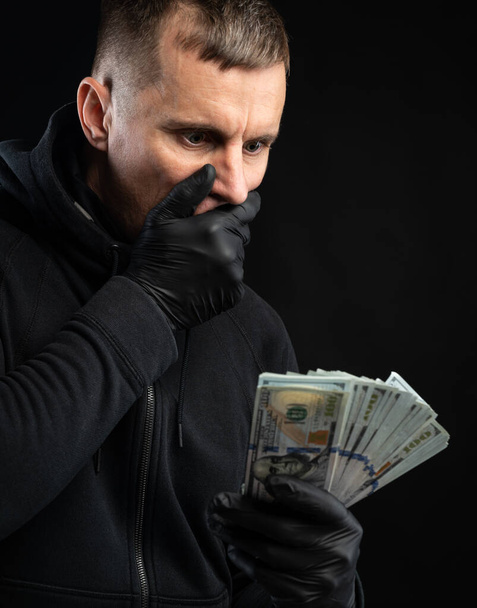 Επιχειρηματική και οικονομική έννοια. Σοκαρισμένος άντρας με μαύρη κουκούλα κρατώντας ένα πακέτο αμερικάνικα δολάρια πάνω από μαύρο φόντο. - Φωτογραφία, εικόνα