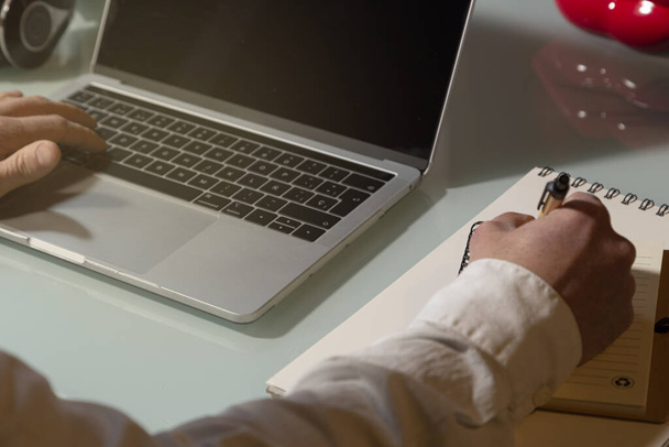 Egy üzletember kezei, aki laptopot használ, és jegyzetel otthonról. Jegyzetfüzet és toll az asztalon. Telework koncepció - Fotó, kép