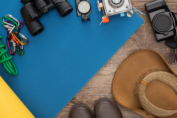 Draufsicht auf Wanderausrüstung auf blauer Isomatte, Fotokamera, Stiefel und Hut auf Holzoberfläche - Foto, Bild