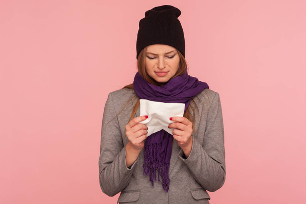 Portrait of depressed flu-sick woman in warm hat and scarf holding tissue, suffering runny nose, sneeze and high temperature, influenza symptoms. indoor studio shot isolated on pink background - 写真・画像