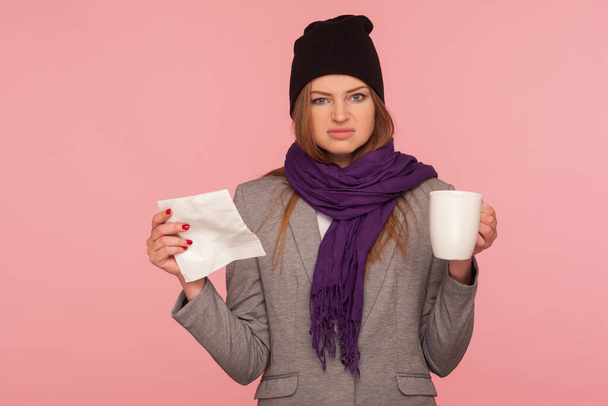 Portrait of irritated flu-sick woman in warm hat and scarf holding tissue and cup of hot beverage, looking with annoyed expression, treating influenza. indoor studio shot isolated on pink background - Фото, изображение