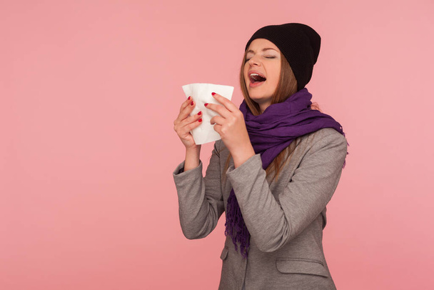 Influenza epidemic. Portrait of flu-sick woman in warm hat and scarf sneezing hard in napkin, blowing nose, suffering allergy symptom, feeling unhealthy. indoor studio shot isolated on pink background - Photo, image