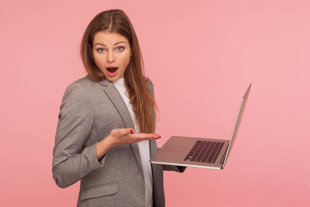 Portret van een verbijsterde zakenvrouw die een laptop vasthoudt en met een verbaasd gezicht naar de camera kijkt, computer gebruikt om te studeren, online werkt, geschokt door de toepassing. studio schot geïsoleerd op roze achtergrond - Foto, afbeelding