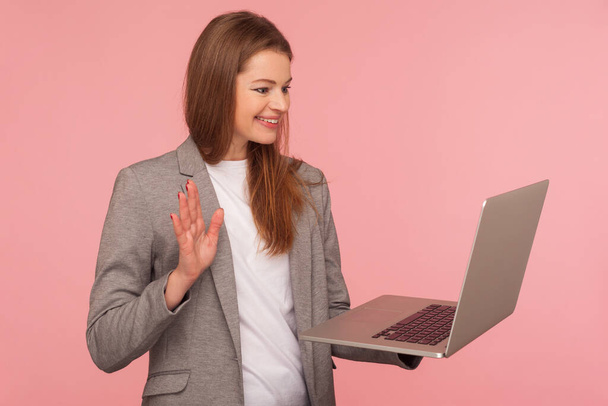 Portrait of positive businesswoman in elegant suit jacket talking on video call with hello hand gesture, welcoming on online conference, using laptop for communication. indoor studio shot isolated - Foto, Bild