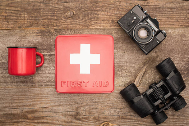 top view of first aid kit, photo camera, binoculars and red metal cup on wooden table - Fotografie, Obrázek