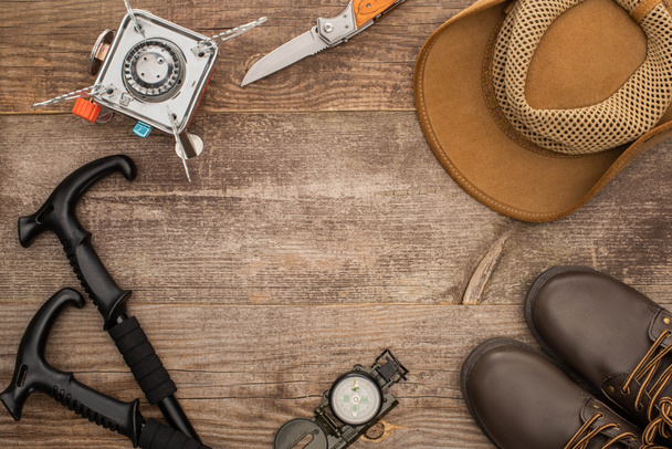 Draufsicht auf Wanderstöcke, Gasbrenner, Jackenmesser, Hut, Stiefel und Kompass auf Holztisch - Foto, Bild