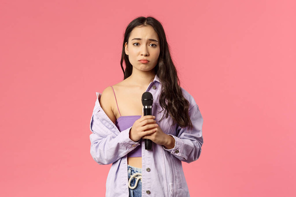 Portré komor és buta aranyos ázsiai lány végre szomorú dalt a közönség előtt, énekel karaoke, duzzogás és nézd magányos kamera, gazdaság mikrofon, zavarban énekelni, rózsaszín háttér - Fotó, kép