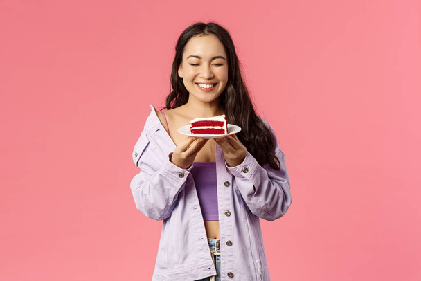 Портрет счастливой, улыбающейся и беззаботной азиатской девушки закрыть глаза и чувствовать себя счастливым держа любимый десерт, есть вкусный торт, падение диеты и наслаждаться сладостями, стоя розовый фон
 - Фото, изображение