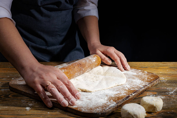 Female hands knead the dough on a wooden antique table on a dark background, close-up, shallow depth of field, beautiful directional lighting. Concept of home baking and comfort. - Fotoğraf, Görsel