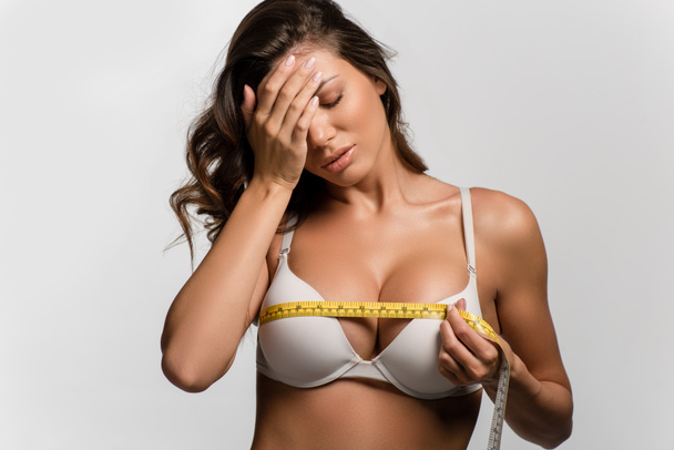 displeased girl holding hand on face while measuring breasts with measuring tape isolated on white - Photo, Image