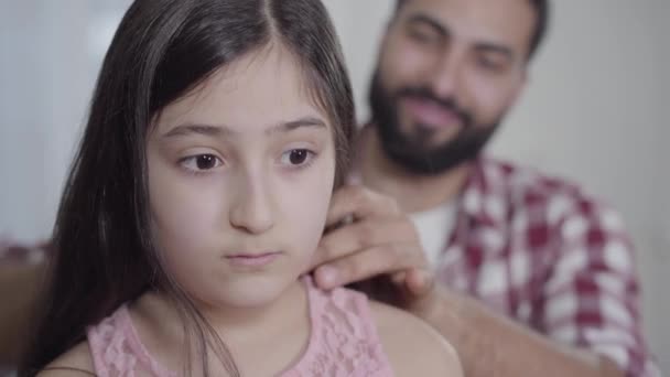 Close-up face of beautiful little girl with brown eyes standing indoors as blurred man combing her long black hair at the background. Middle Eastern father taking care of pretty daughter. - Séquence, vidéo