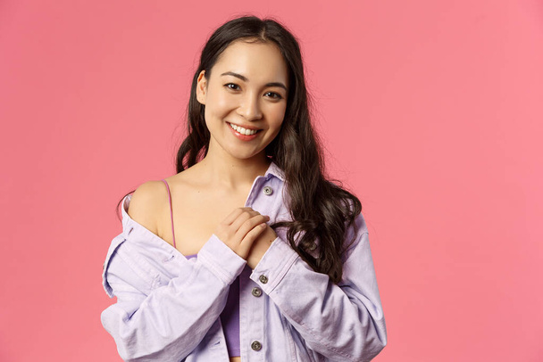 Close-up portrait of tender, romantic asian girl keep loving dearest memories in her heart, press hands to chest, smiling and looking thankful camera, standing pink background - Photo, Image