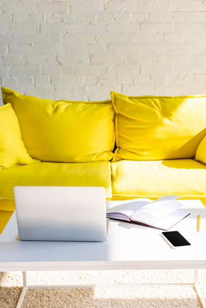 Sarı kanepeli oturma odası, dizüstü bilgisayarlı masa, akıllı telefon ve güneş ışığında not defteri. - Fotoğraf, Görsel