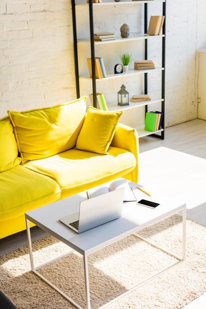 sala de estar com sofá amarelo, prateleira e mesa com laptop, smartphone e bloco de notas em luz solar
 - Foto, Imagem