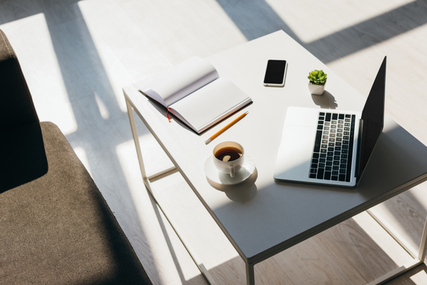 leerer Arbeitsplatz mit Laptop, Smartphone, Notizblock und Kaffee im Sonnenlicht - Foto, Bild