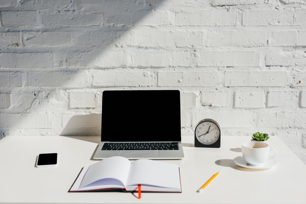 domácí kancelář s notebookem, smartphonem, prázdným notepadem, hodinami a šálkem kávy na slunci - Fotografie, Obrázek