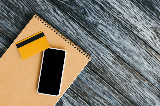 vista superior del bloc de notas, tarjeta de crédito y teléfono inteligente con pantalla en blanco en la superficie de madera oscura
 - Foto, Imagen