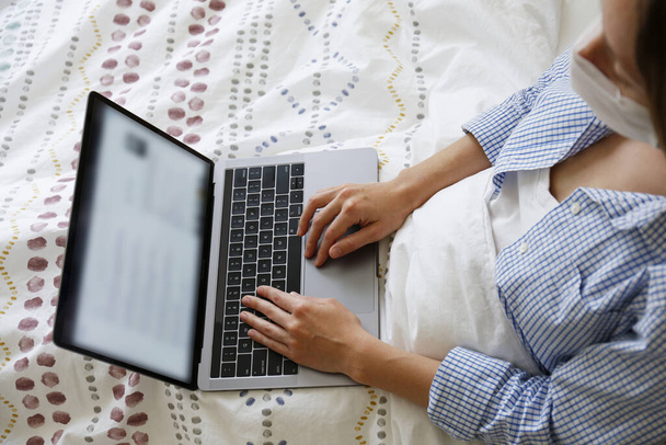 Close up shot of young blonde woman wearing disposable face mask working at home due to coronavirus quarantine concept. Beautiful female sitting in bed with laptop. Background, copy space. - Foto, immagini
