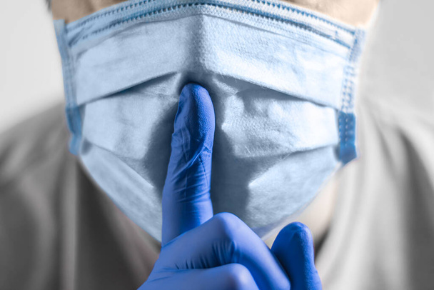 Μια κοντινή φωτογραφία ενός χεριού σε ένα προστατευτικό ιατρικό γάντι δείχνει μια χειρονομία σιωπής ενάντια σε ένα πρόσωπο σε μια προστατευτική μάσκα - Φωτογραφία, εικόνα