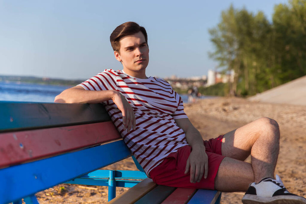 A handsome, young guy, a man in a white striped shirt and shorts is sitting on a bench by the river. The beach at the resort. Breathe the sea air, iodine deficiency. - Foto, afbeelding