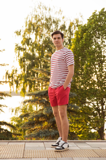 A handsome, young guy, a man in a white striped shirt and shorts stands against a background of trees. in the park. Walk in the fresh air. To go on the road. Model posing. Million, generation z. - Foto, afbeelding