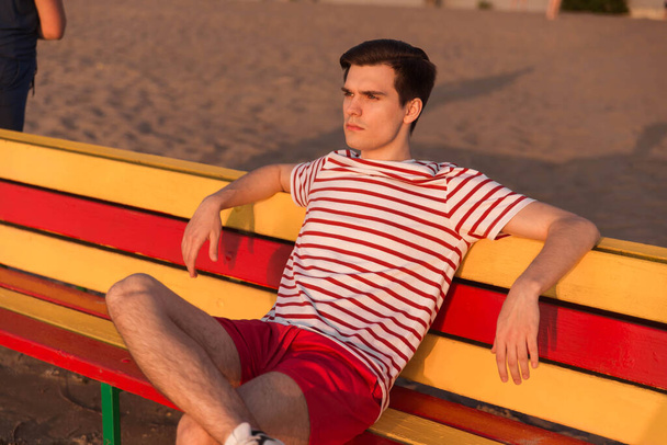 A handsome, young guy, a man in a white striped shirt and shorts is sitting on a bench by the river. The beach at the resort. Breathe the sea air, iodine deficiency. - Foto, Imagem