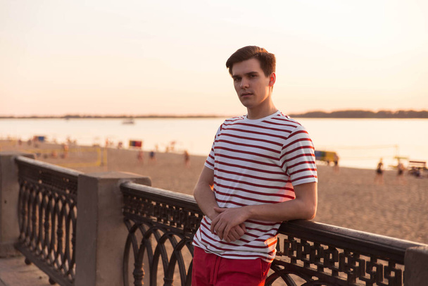 A handsome, young guy, a man in a white striped shirt is leaning on a bridge and is looking into the distance. Embankment, beach resort. Life style. Character from old movies. - Photo, image