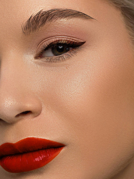 Close-up beauty of half female face with creative fashion evening make-up. Black arrows on the eyes and extremely long eyelashes, on full red lips matte scarlet lip color. Well-groomed skin after spa - Фото, изображение