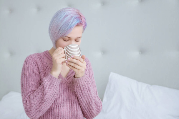 A beautiful young woman with blue hair is sitting on a bed in a bedroom dressed in a lilac, purple sweater, holding a mug with hot coffee or tea and drinking it. Copy space. - Foto, Imagen