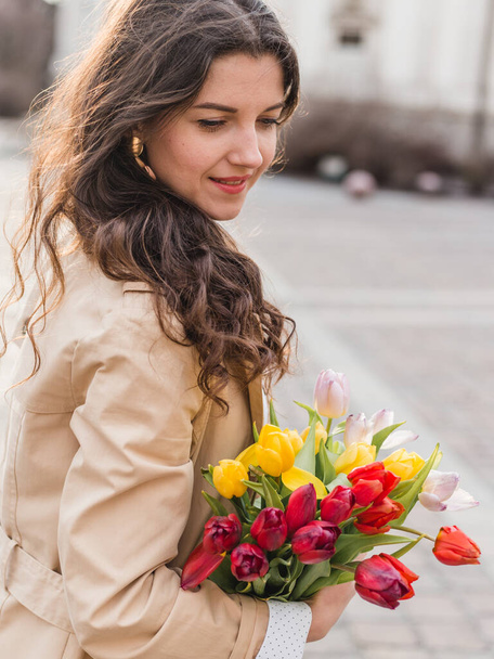 Beautiful young woman with spring tulips flowers bouquet at city street. Happy girl walking outdoors. Spring portrait of pretty female in park. - Foto, Imagem