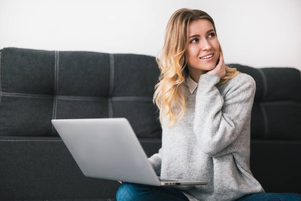 young blond beautiful woman sits on the sofa working from home on her laptop during quarantine looks happy, cozy home atmosphere concept. - Photo, Image