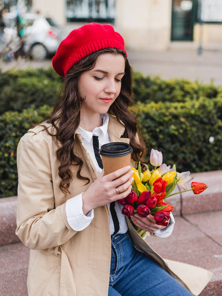 Beautiful young woman with coffee to go and spring tulips flowers bouquet at city street. Happy girl walking outdoors. Spring portrait of pretty female in park. - Photo, Image