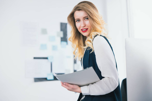 young woman manager in smart suit stands in her modern office with documents in her hands, looks concentrated while studying business contracts, multitasking, work concept. - Foto, Bild