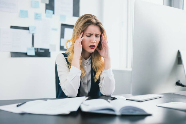 νέα γυναίκα διευθυντής εργάζεται στο σύγχρονο γραφείο της, φαίνεται κουρασμένος που πάσχουν από ξαφνικό πονοκέφαλο, multitasking, άγχος έννοια της εργασίας. - Φωτογραφία, εικόνα