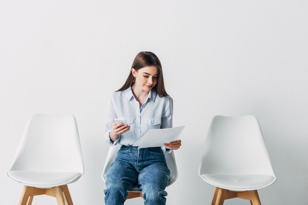 Beautiful woman using smartphone and holding resume on chair in office  - Foto, immagini