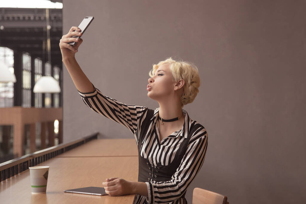 Attractive blonde woman sits at the table and makes selfie, smile, picture. Happy girl with coffee. Take pictures of yourself in a cafe. Duck face, lips with a bow. - Foto, Imagen