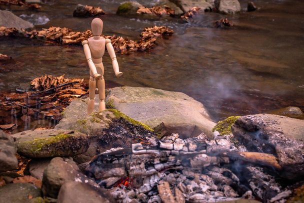 Un hombrecito articulado se calienta por el fuego a orillas de un río de montaña
 - Foto, Imagen