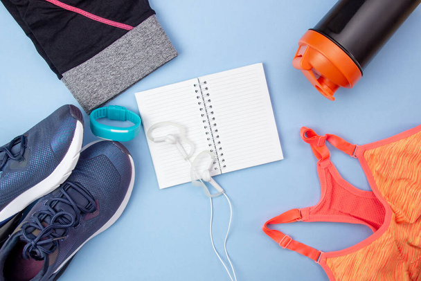 Sports flat layout. Fitness accessories sneakers, headphones, clothes, bottle, watch and an open notebook for writing on a blue background. Sports Planning. - Zdjęcie, obraz