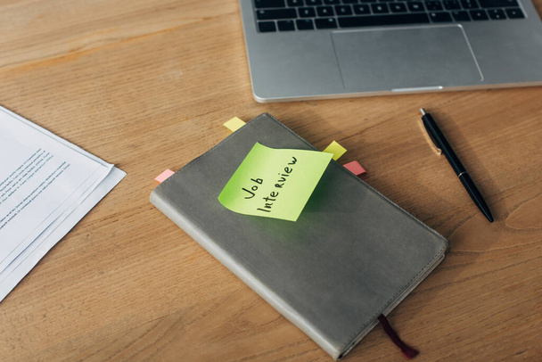 Notebook met sollicitatiegesprek belettering op plakkerige notitie in de buurt van laptop en documenten op tafel - Foto, afbeelding