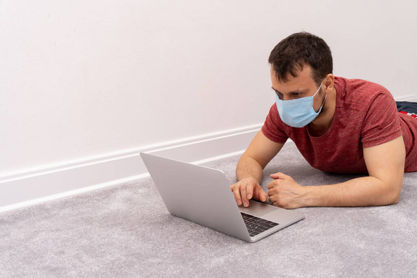 Man in facial protective medical mask lay on floor and work at home at laptop - Foto, Bild