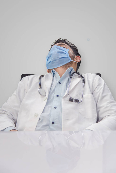 pandemia do Coronavirus COVID-19. Cansado, médico exausto de uniforme está dormindo na mesa do hospital ou clínica após o dever duro. macho com máscara e estetoscópio ao redor do pescoço e olhos fechados
 - Foto, Imagem