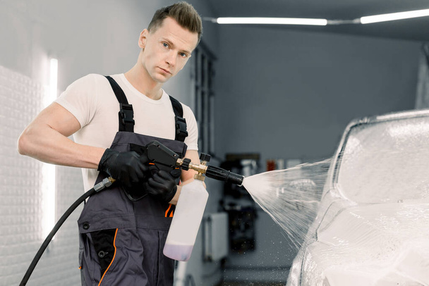Zbliżenie portret pracownika myjni samochodowej, Hansome młody człowiek w kombinezonach ochronnych, czyszczenie samochodu z wysokociśnieniowym strumieniem wody w myjni samochodowej, spryskiwanie pianki czyszczącej - Zdjęcie, obraz