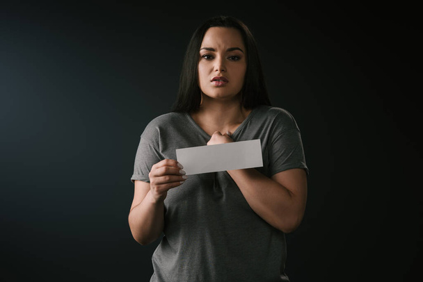 Front view of beautiful plus size girl confused and holding empty card on black background - Photo, image
