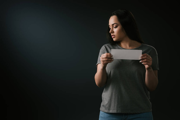 Přední pohled smutné plus velikost dívka drží prázdnou kartu na černém pozadí - Fotografie, Obrázek