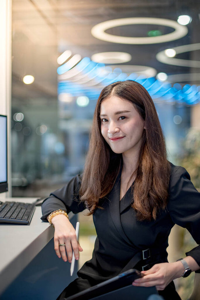 Концепция работающих женщин. Привлекательная молодая азиатская предпринимательница, использующая цифровой планшет и компьютер, работающая в современном офисе. Уверенная женщина-предприниматель улыбается на работе. Работа с технологиями
. - Фото, изображение