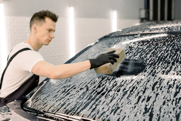 Jovem lavador de carros do sexo masculino limpando o pára-brisas do carro com a ajuda de foem especial e esponja amarela. Lavar o carro à mão, esponja e bolhas de espuma
 - Foto, Imagem