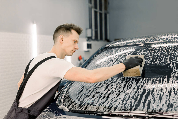 Car wash and clean with shampoo and sponge. Young handsome man worker washing soapy car windshield with sponge on a car wash. - Фото, изображение