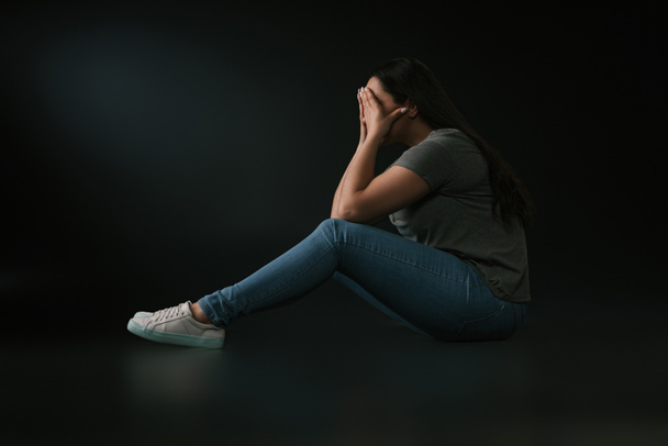 Side view of crying plus size girl covering face with hands and sitting on black background - Foto, Imagen